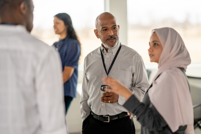 A small group of business professionals stand around in a conference room as they casually meet to discuss business strategies.  They are each dressed semi-casually and have neutral expressions on tehir faces.